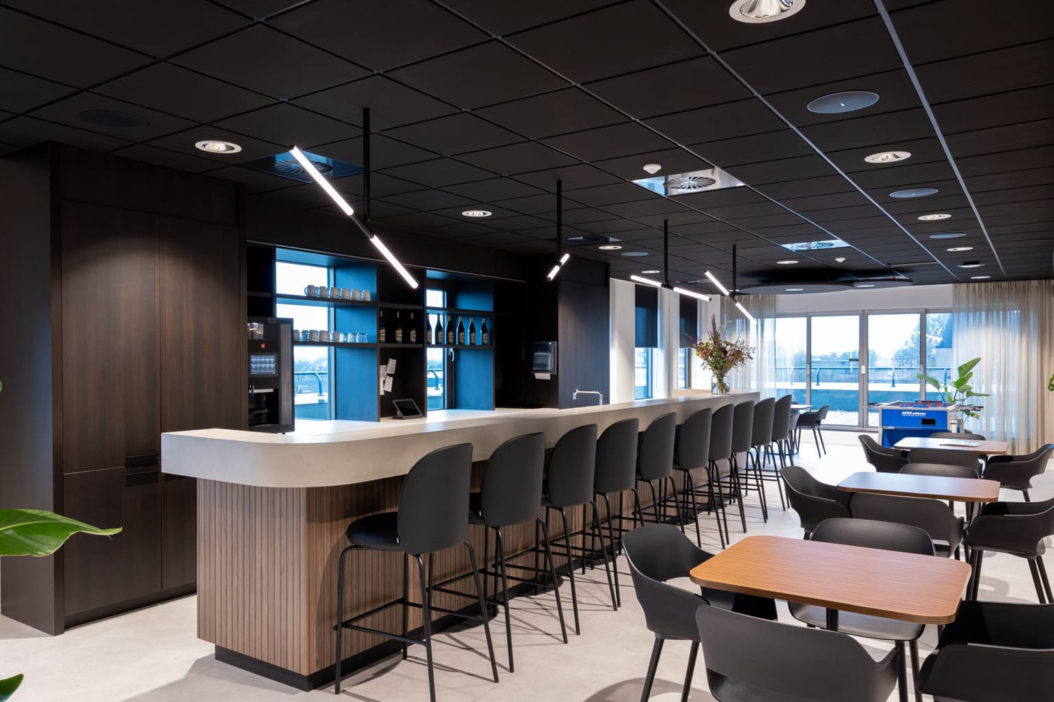 Bar in een kantine met een donkerhouten poot en een licht blad. Langs de bar staan kantinestoelen.