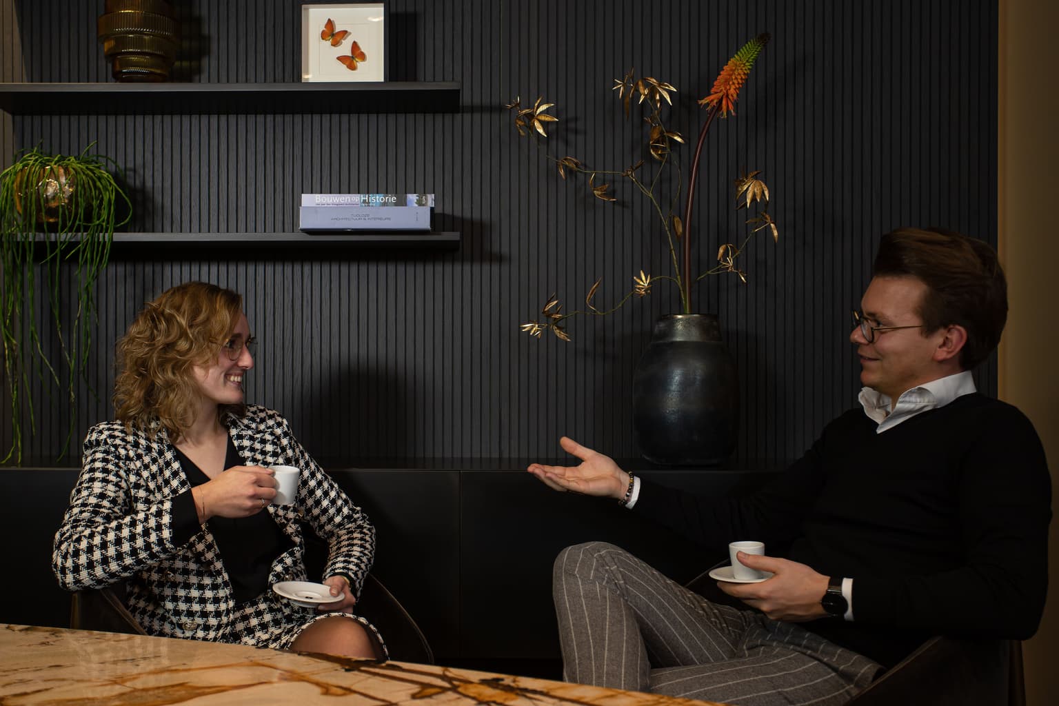 Twee mensen zitten aan een tafel. Links zit een vrouw met een geblokte blazer, dit is Andrea Ebbers. Rechts zit een man die naar haar gebaard en een bril opheeft. Dit is Herwin Ebbers. 